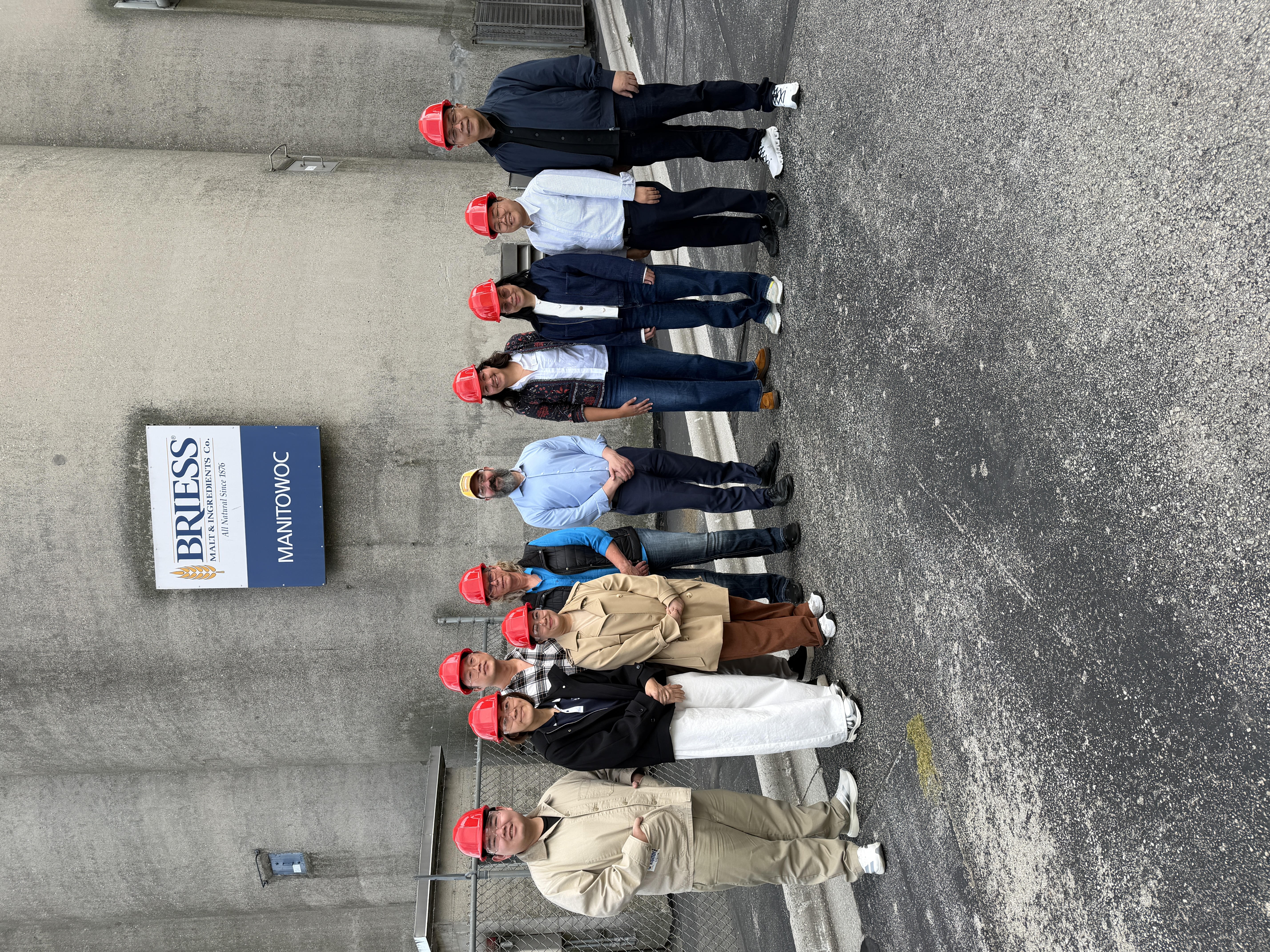 Mission members and IABC staff pose in hard hats in front of the Briess sign in Manitowoc.
