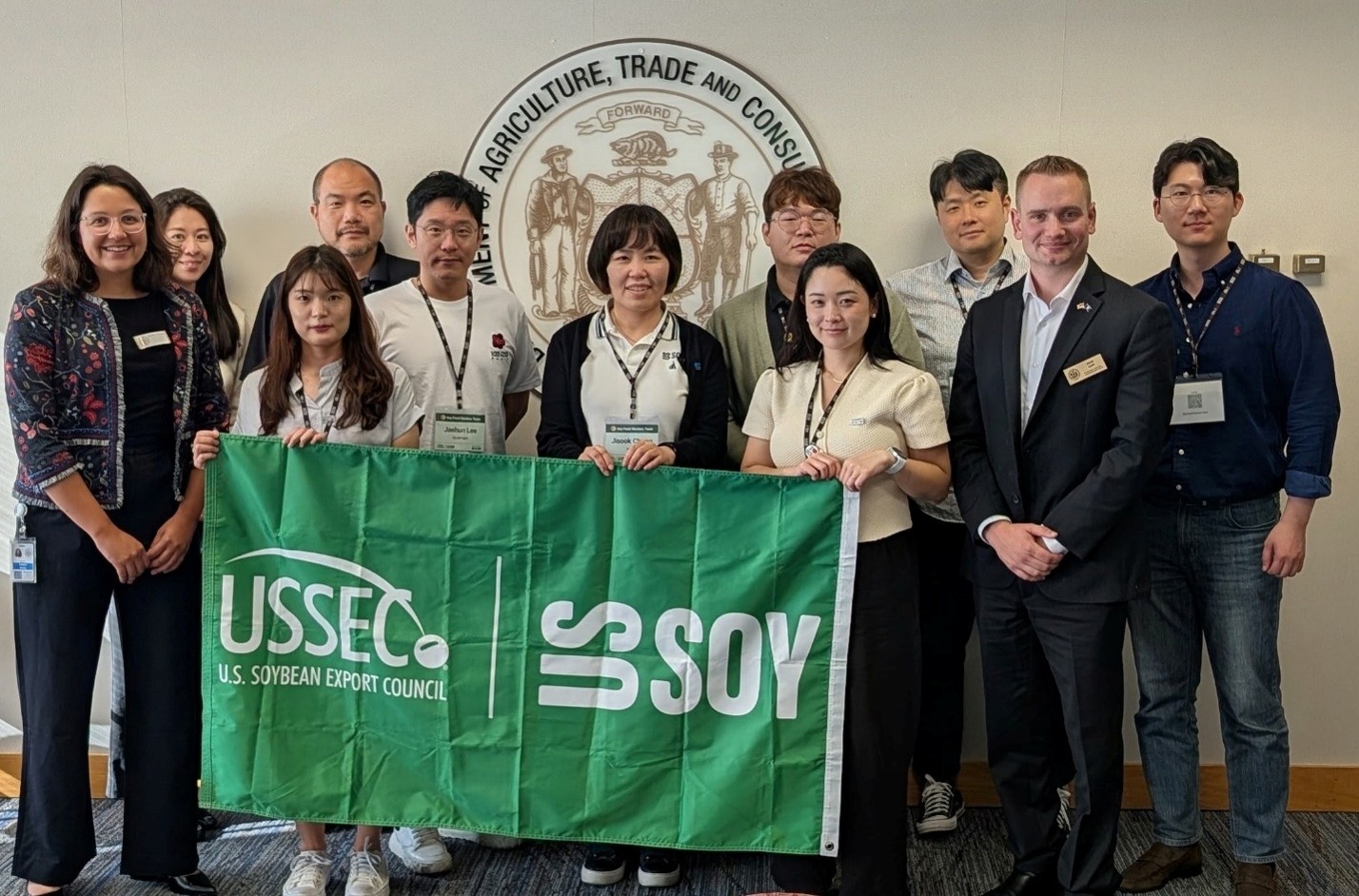 Korean delegates and IABc staff pose in front of DATCP logo and hold sign saying "USSEC" and "US Soy".