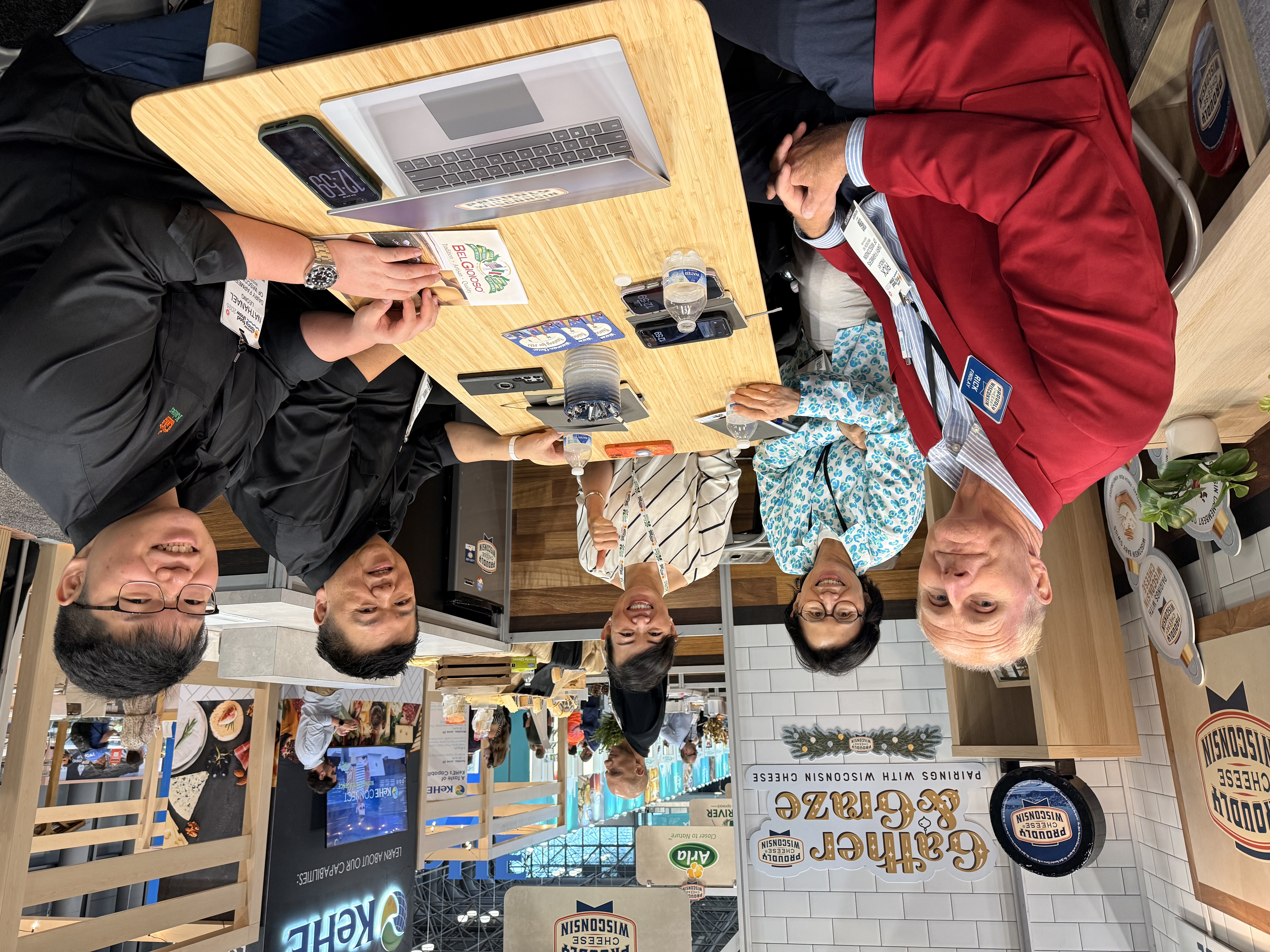 Five people sit around a table with a laptop, phones, and notebooks, with an exposition and booths in the background.
