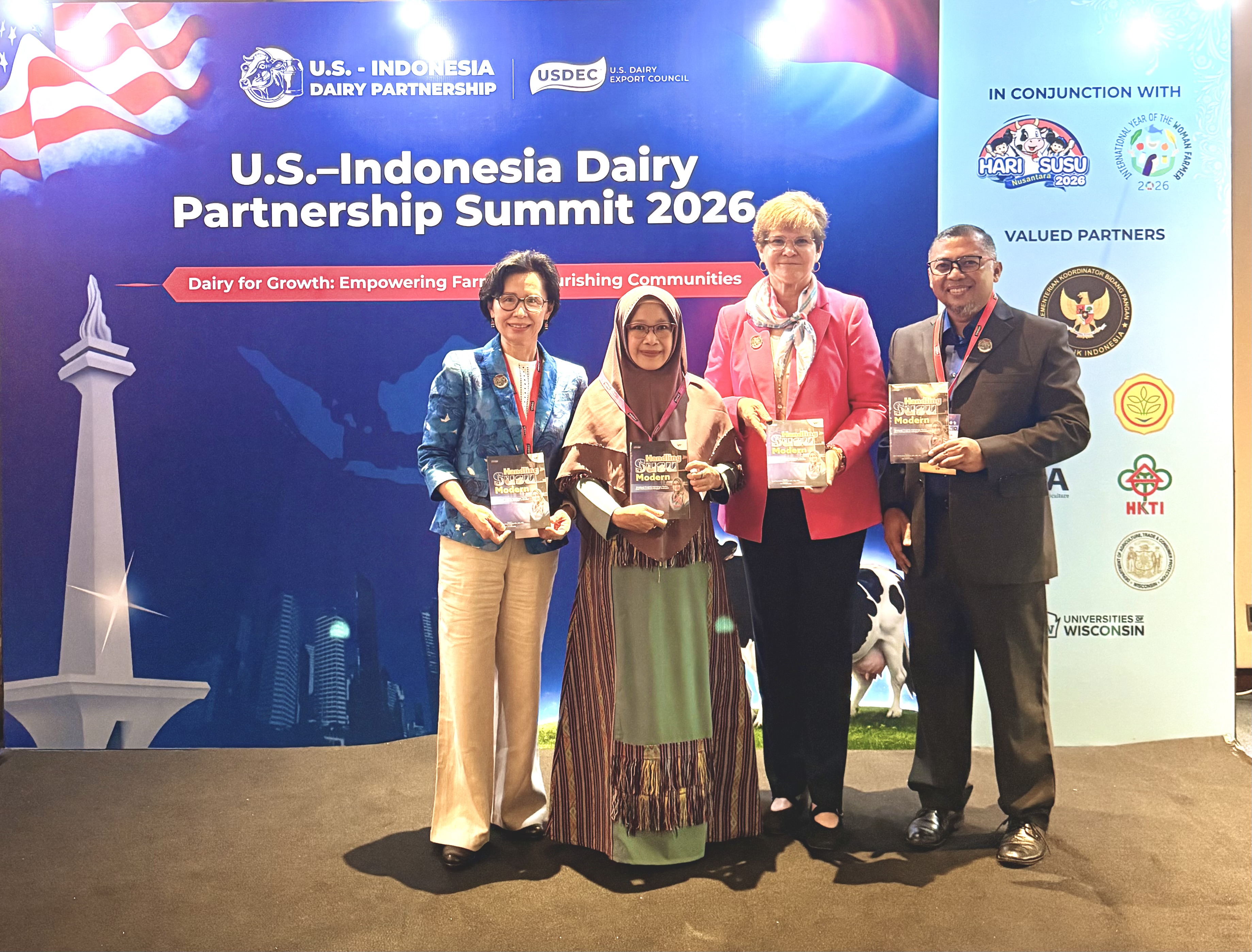 A group of four people smile and hold books in front of a colorful backdrop that says "USIDP Summit 2026".