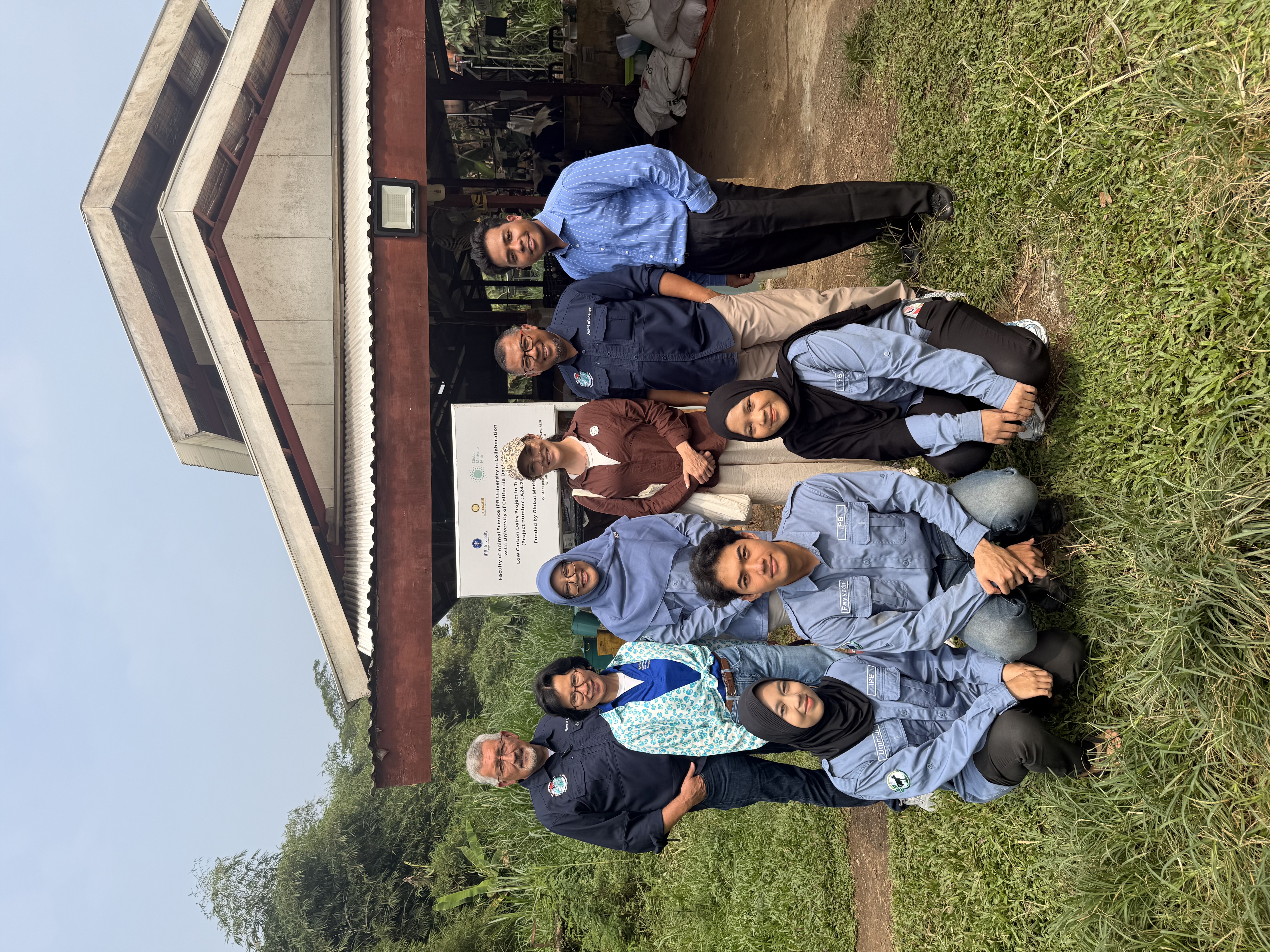 A group of nine people smile in front of an outdoor structure with lots of surrounding vegetation and trees.