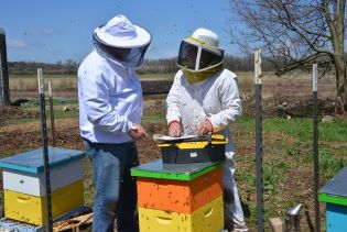 Two individuals talk outside  while standing beside beekeeping equipment. 